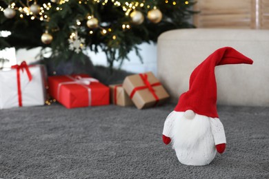Photo of Cute Scandinavian gnome on carpet near Christmas tree and gift boxes in room. Space for text