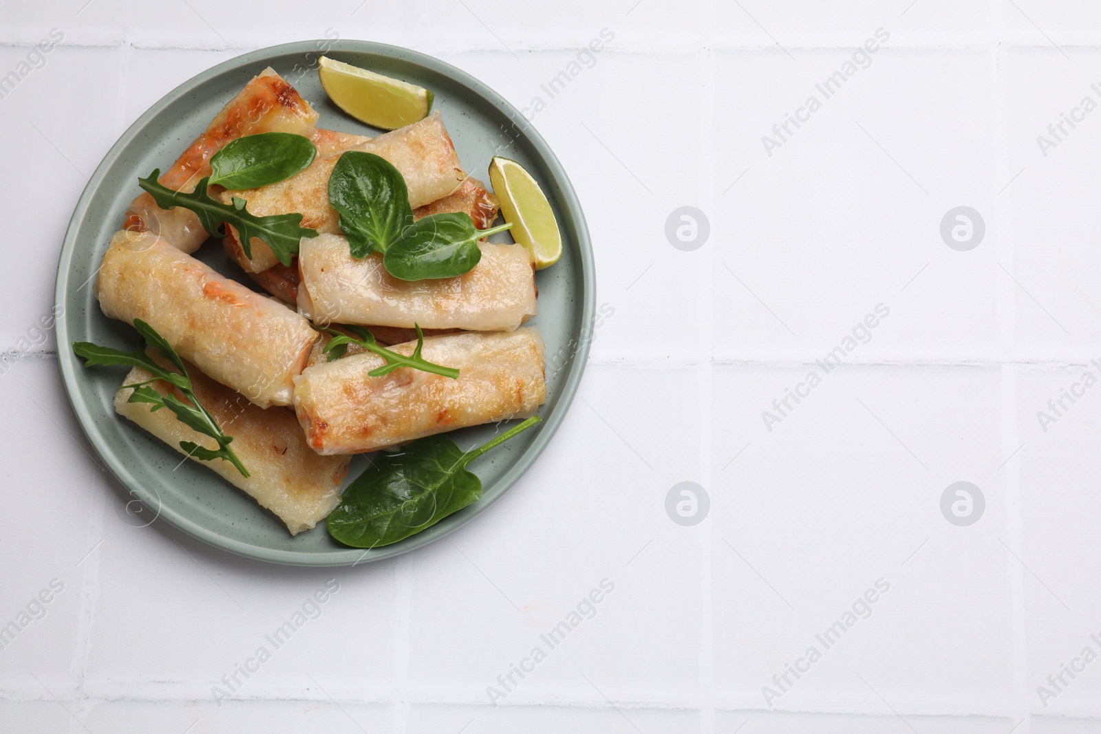 Photo of Plate with tasty fried spring rolls, spinach, arugula and lime on white tiled table, top view. Space for text