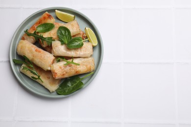 Plate with tasty fried spring rolls, spinach, arugula and lime on white tiled table, top view. Space for text