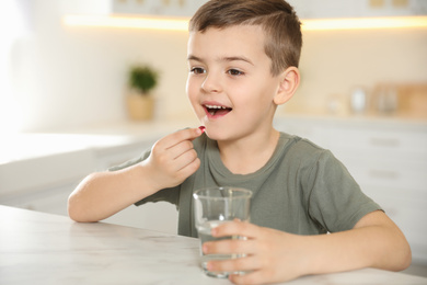 Photo of Little boy with glass of water taking vitamin capsule in kitchen