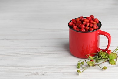 Fresh wild strawberries in mug and green stems on white wooden table. Space for text