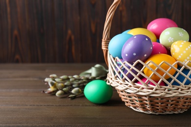 Photo of Colorful Easter eggs in wicker basket and willow branches on wooden background, closeup. Space for text