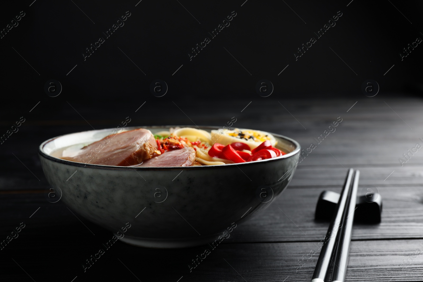 Photo of Delicious ramen in bowl and chopsticks on black wooden table, closeup. Noodle soup