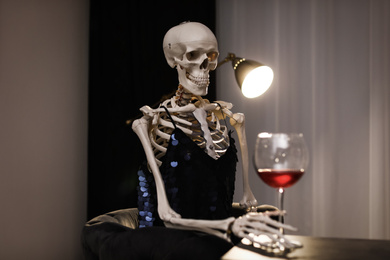 Skeleton in dress with wine sitting at table indoors