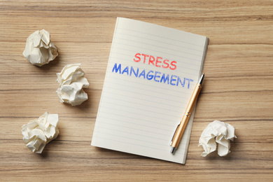 Notebook with text STRESS MANAGEMENT and crumpled paper balls on wooden table, flat lay