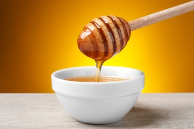 Pouring honey from dipper into bowl at wooden table against golden background, closeup