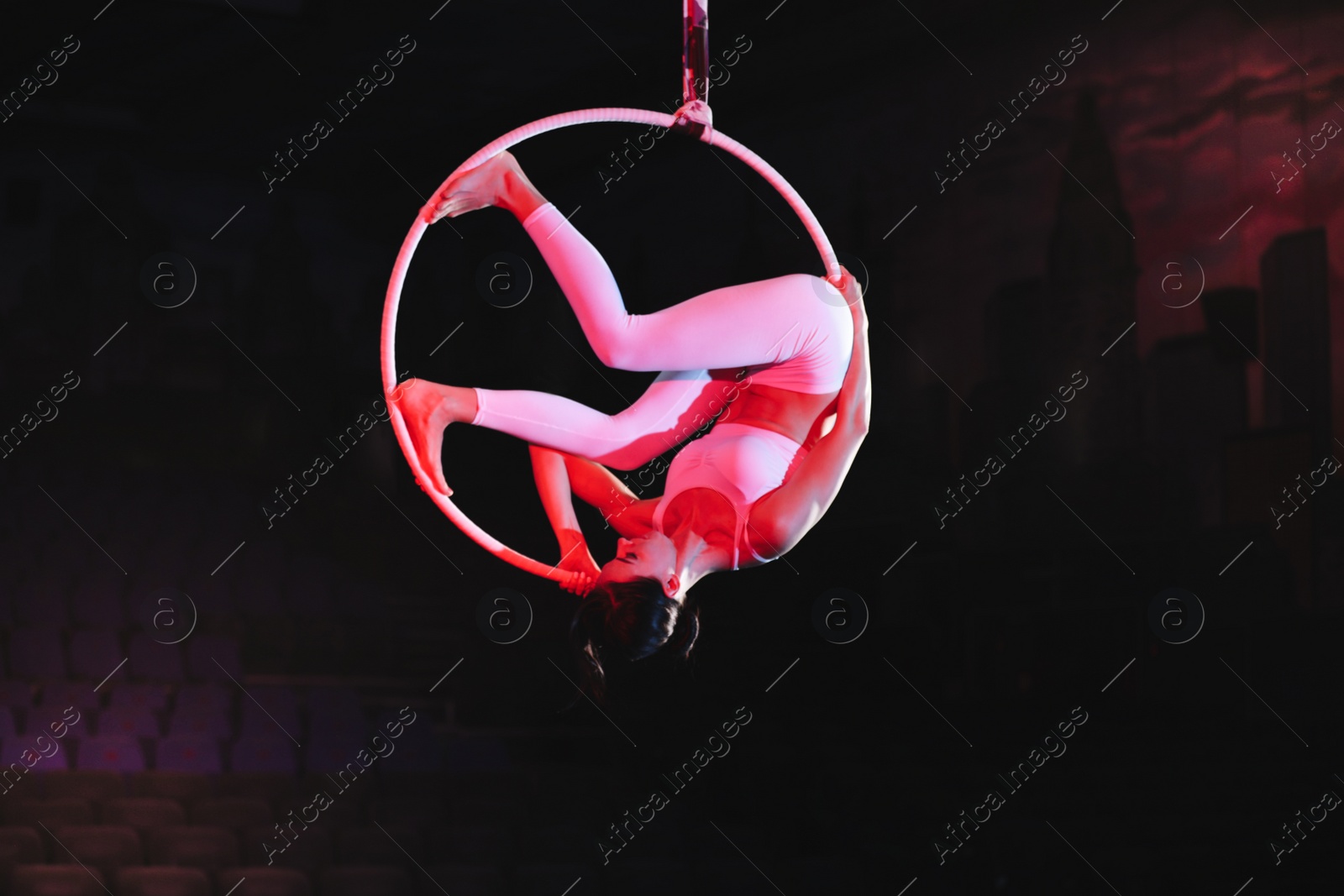 Photo of Young woman performing acrobatic element on aerial ring against dark background