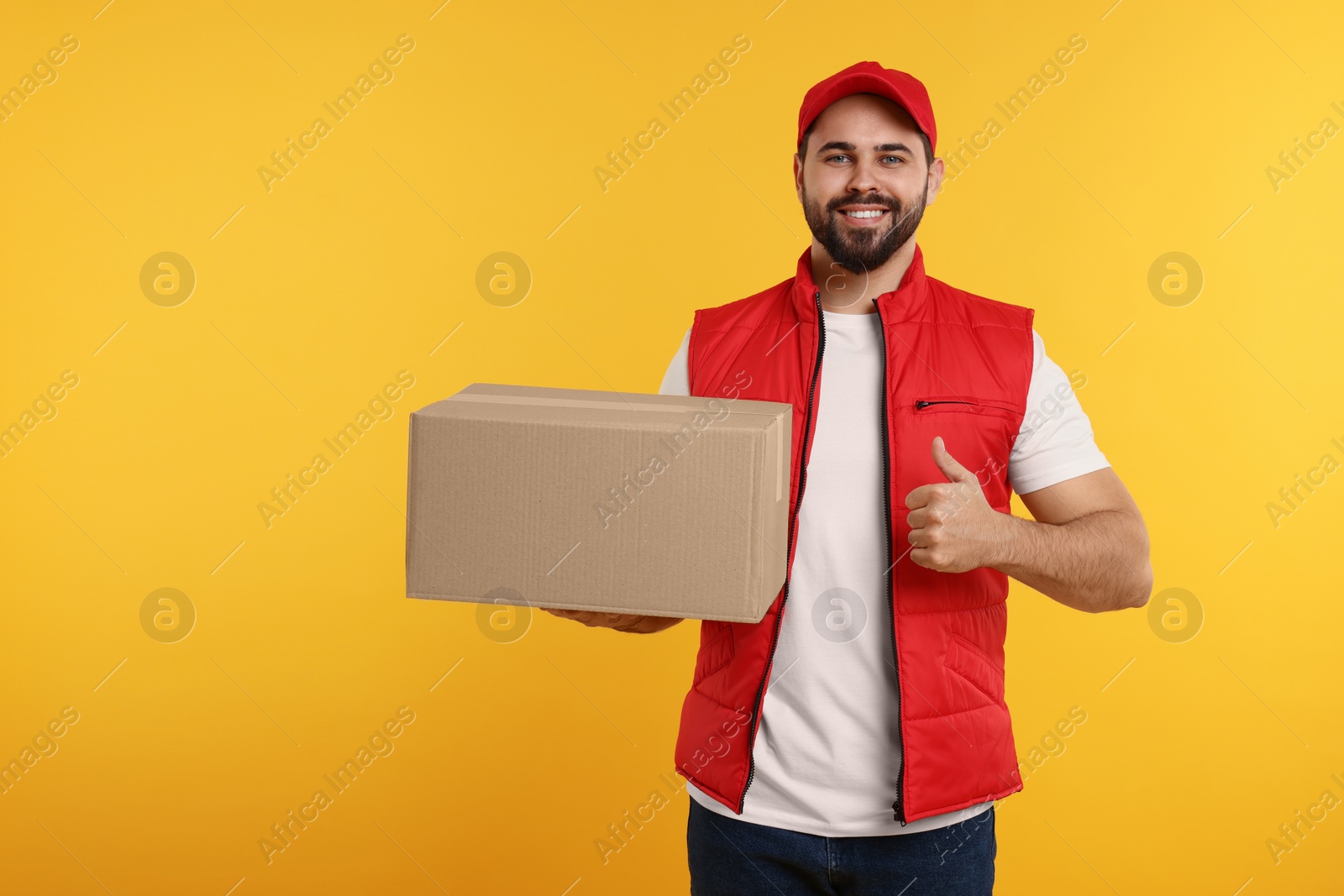 Photo of Happy young courier with parcel showing thumb up on orange background, space for text