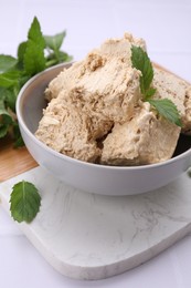 Bowl with pieces of tasty halva and mint leaves on white table, closeup