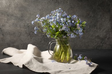 Bouquet of beautiful forget-me-not flowers in glass jug and cloth on gray table