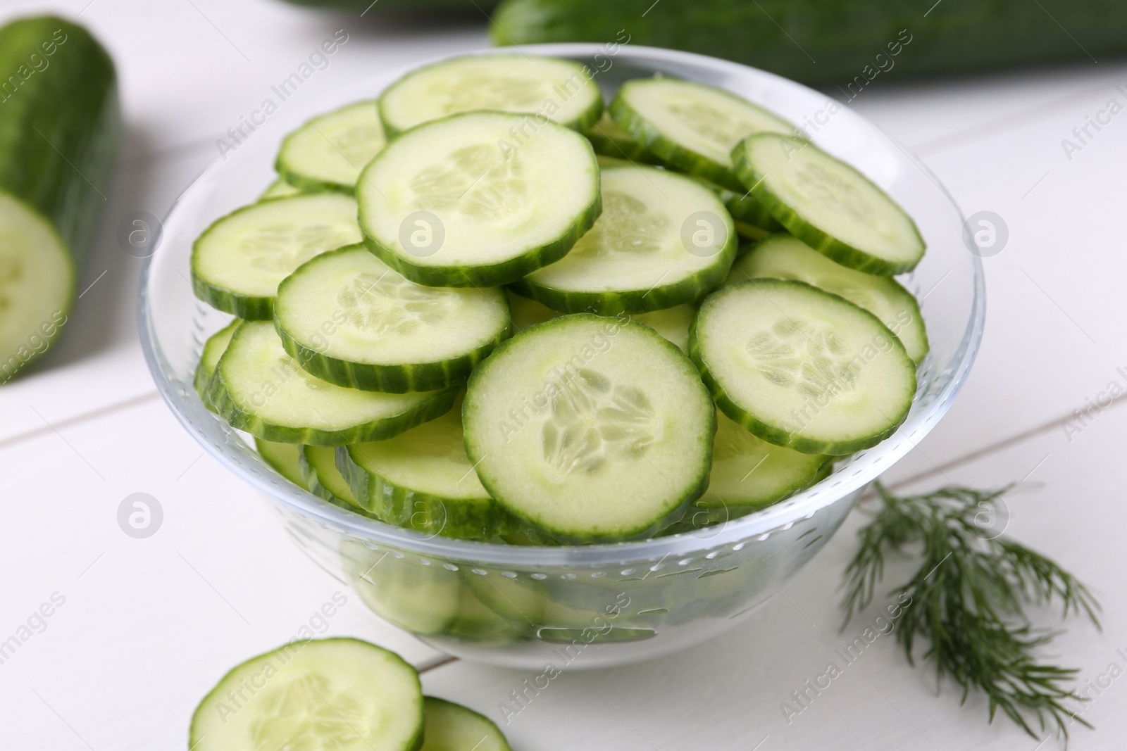 Photo of Cut cucumber in glass bowl and dill on white wooden table, closeup