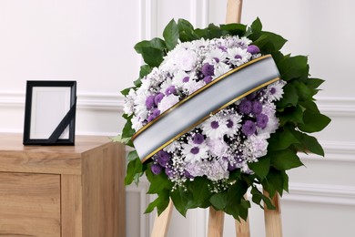 Image of Wreath of violet and white flowers, photo frame with black ribbon in room. Funeral attributes