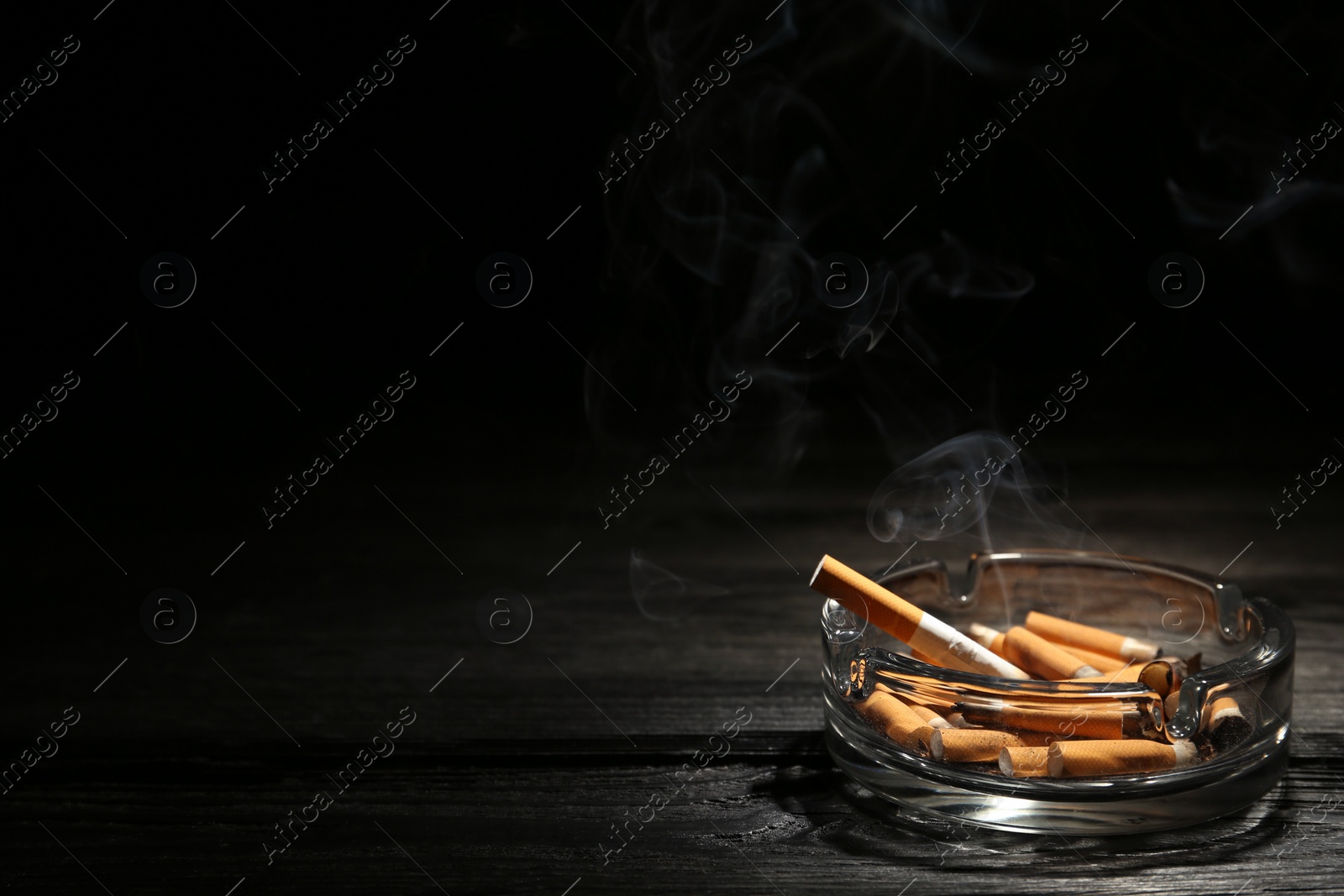 Photo of Smoldering cigarette in glass ashtray on dark wooden table against black background. Space for text