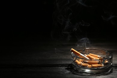 Photo of Smoldering cigarette in glass ashtray on dark wooden table against black background. Space for text