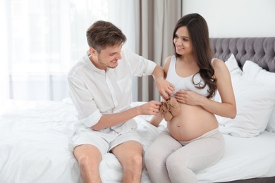 Photo of Young husband touching his pregnant wife's tummy with cute little shoes in bedroom