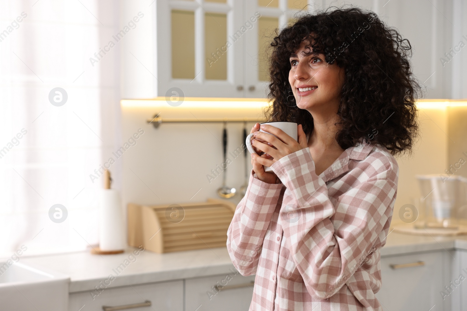 Photo of Beautiful young woman in stylish pyjama with cup of drink in kitchen, space for text