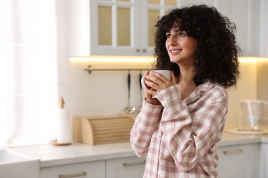 Photo of Beautiful young woman in stylish pyjama with cup of drink in kitchen, space for text