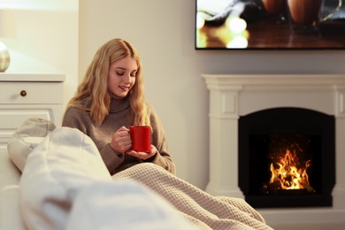 Young woman with cup of hot drink resting near fireplace at home
