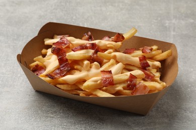 Photo of Tasty potato fries, cheese sauce and bacon in paper container on grey table, closeup