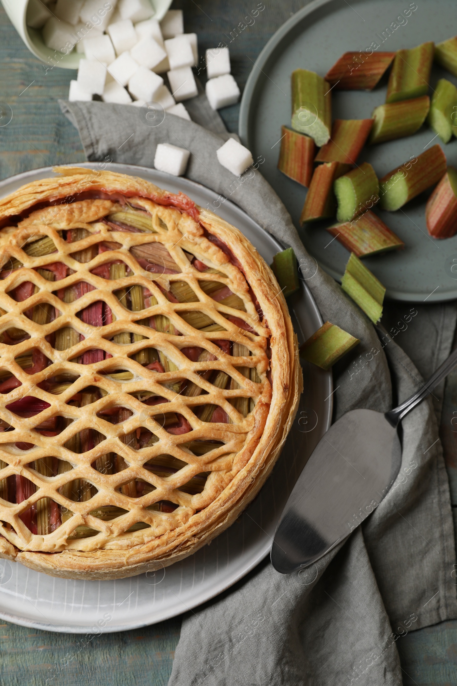 Photo of Freshly baked rhubarb pie, cut stalks, sugar cubes and cake server on wooden table, flat lay