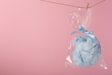 Photo of Packaged sweet light blue cotton candy hanging on clothesline against pink background, space for text