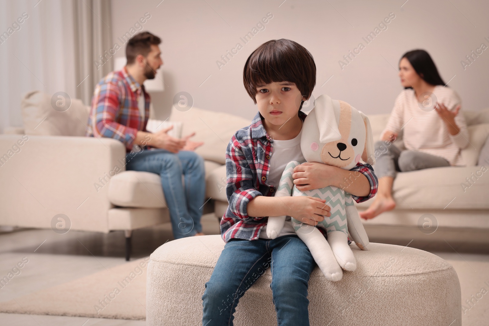 Photo of Sad little boy with toy and his arguing parents on sofa in living room