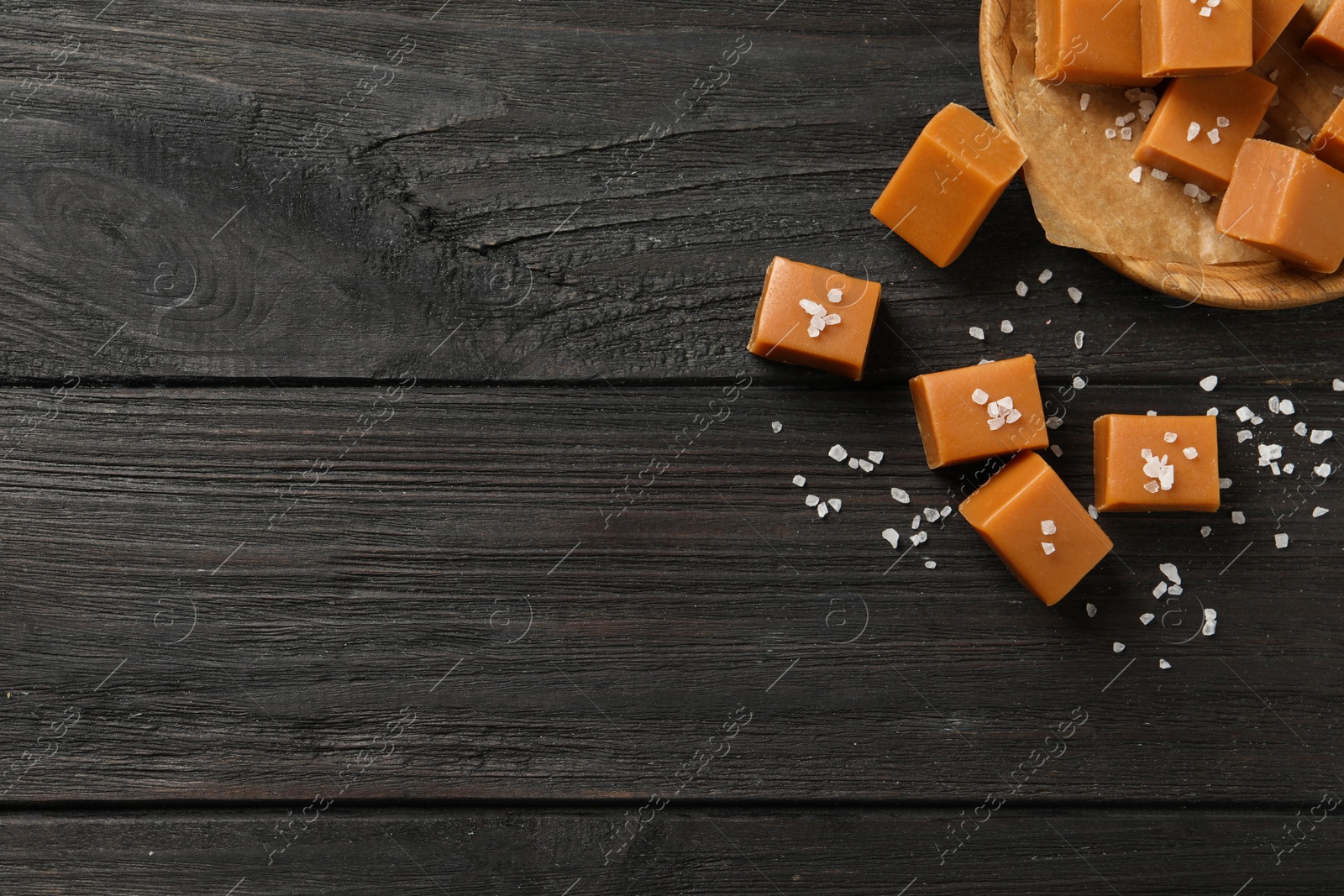 Photo of Yummy caramel candies and sea salt on black wooden table, flat lay. Space for text