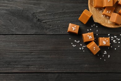 Photo of Yummy caramel candies and sea salt on black wooden table, flat lay. Space for text