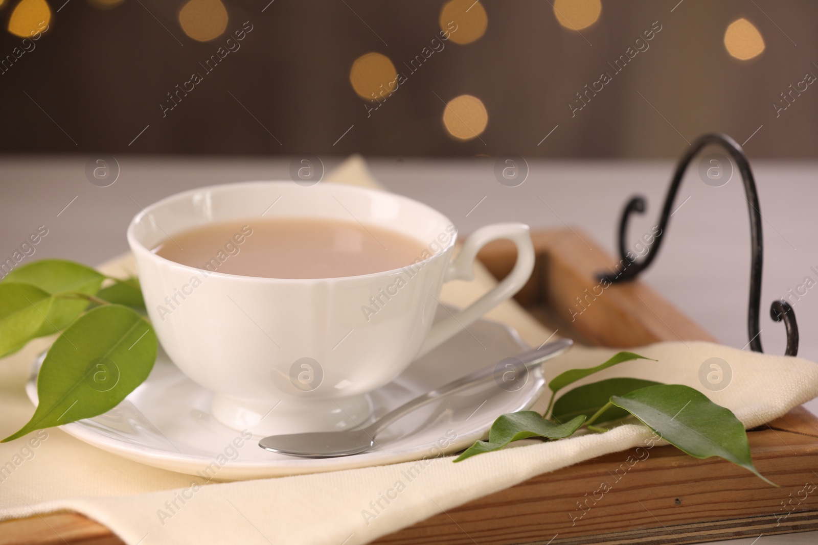 Photo of Cup of aromatic tea with milk, spoon, saucer and green leaves on table, closeup