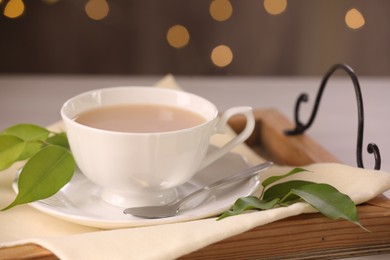 Cup of aromatic tea with milk, spoon, saucer and green leaves on table, closeup