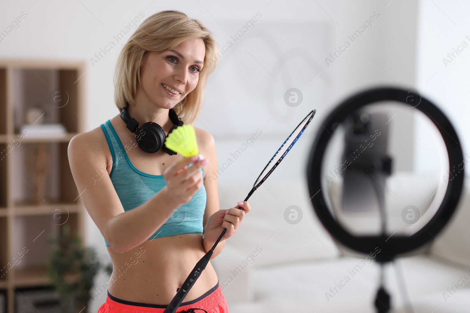 Photo of Smiling sports blogger holding badminton racket and shuttlecock while streaming online fitness lesson with smartphone at home