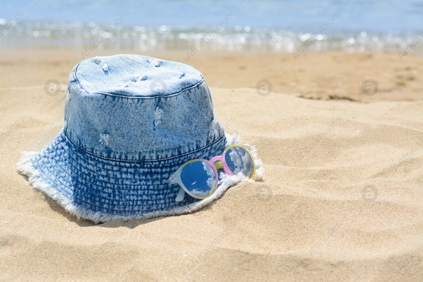 Photo of Jeans hat and sunglasses on sand near sea, space for text. Beach accessories