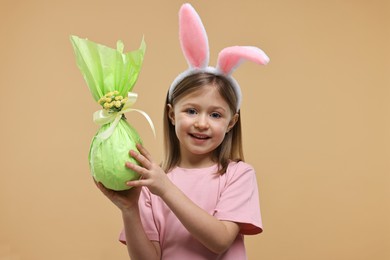 Photo of Easter celebration. Cute girl with bunny ears holding wrapped gift on beige background