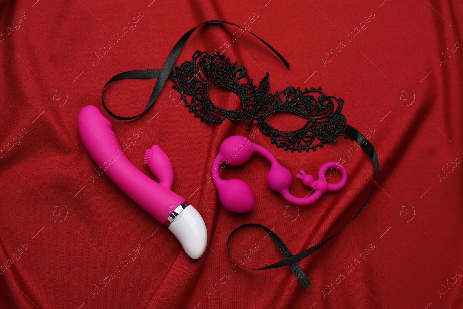 Photo of Pink sex toys and black lace mask on red fabric, flat lay