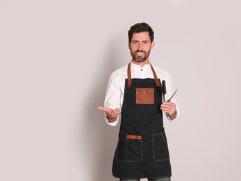 Smiling hairdresser in apron holding scissors and comb on light grey background, space for text