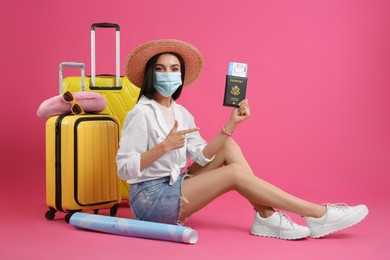 Photo of Female tourist in medical mask with passport, ticket and suitcases on pink background. Travelling during coronavirus pandemic