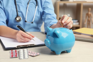 Photo of Doctor putting coins into piggy bank near pills at wooden table in hospital, closeup. Medical insurance