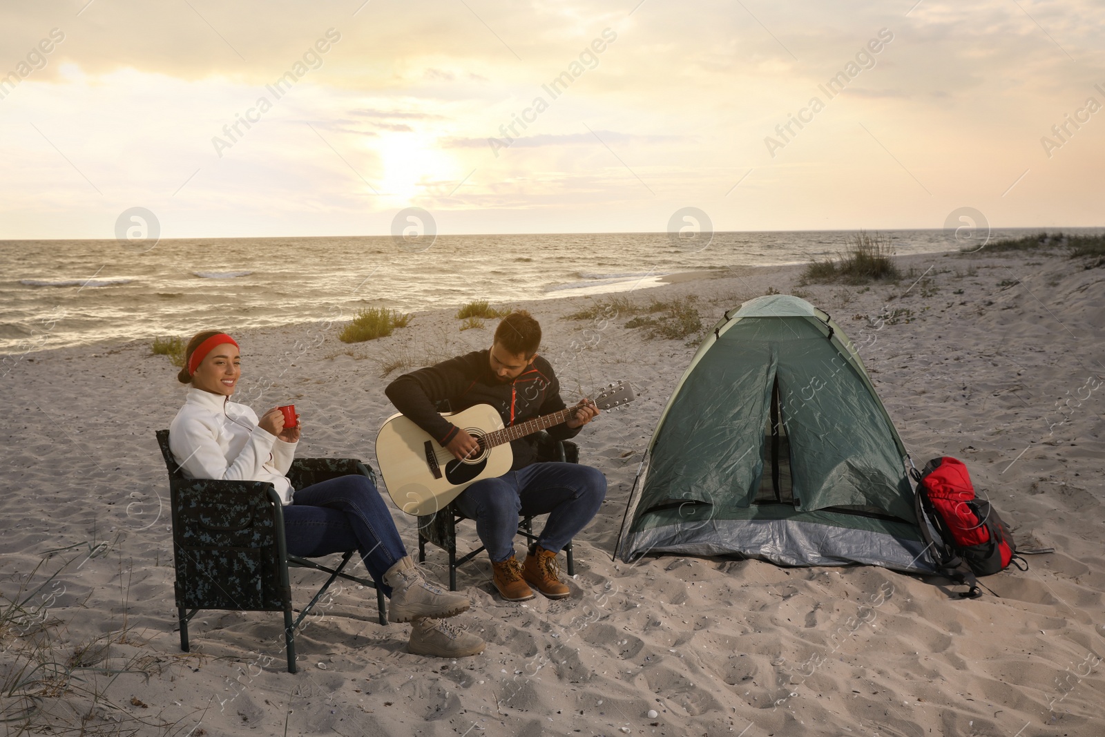 Photo of Young man playing guitar to his beloved girlfriend near camping tent on beach