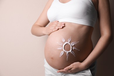 Young pregnant woman with sun protection cream on belly near beige wall, closeup. Space for text