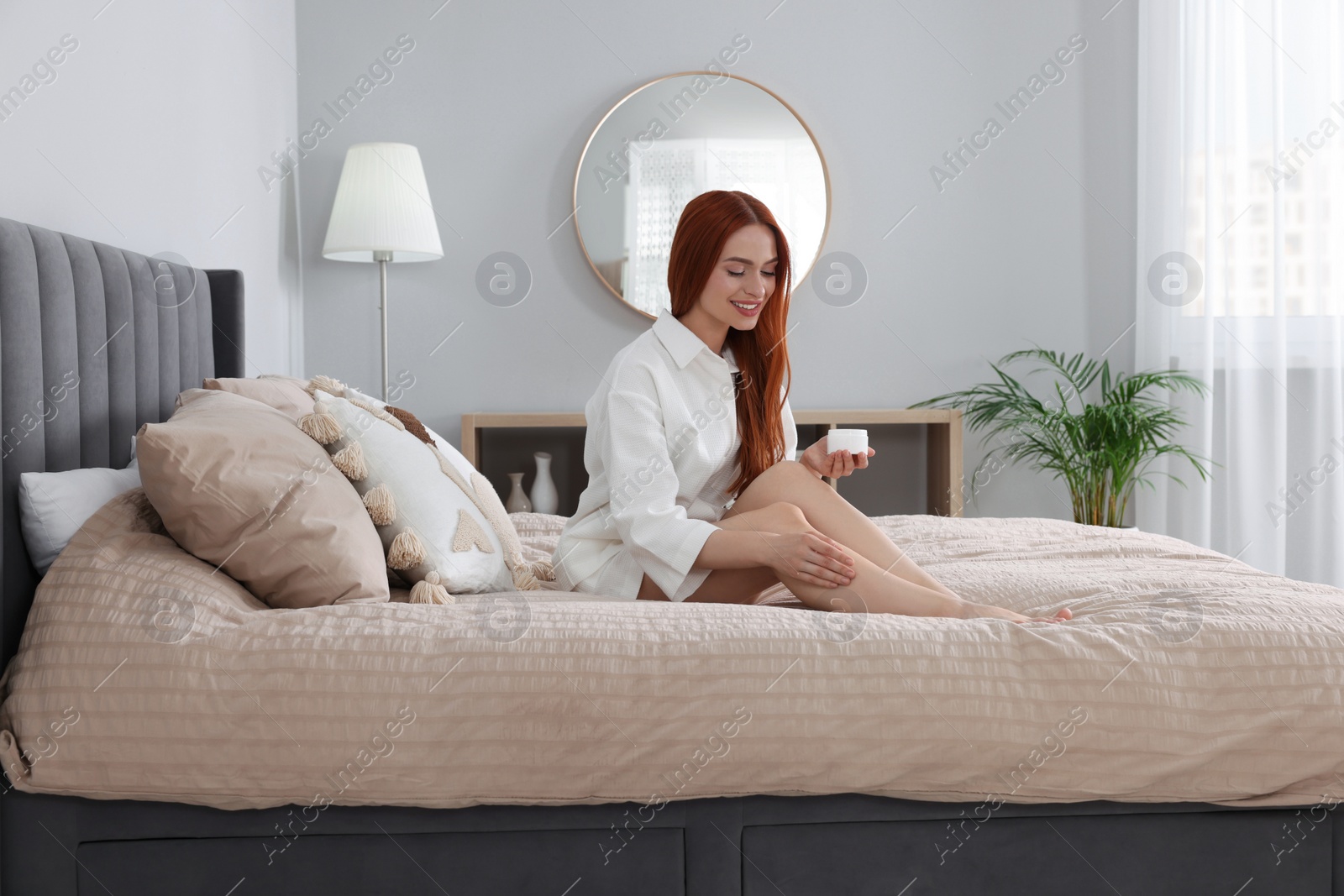 Photo of Beautiful young woman applying body cream onto legs in bedroom