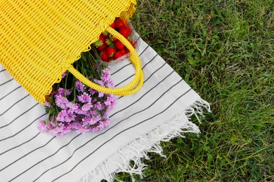 Yellow wicker bag with beautiful flowers and strawberries on picnic blanket outdoors, top view