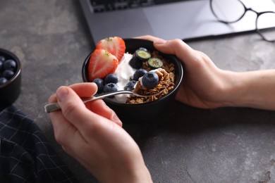 Woman eating tasty granola with yogurt and berries at workplace, closeup