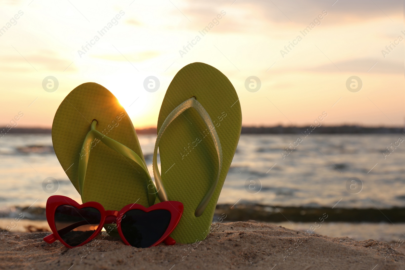 Photo of Stylish flip flops with sunglasses on sand near sea, space for text. Beach accessories