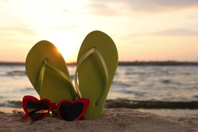 Stylish flip flops with sunglasses on sand near sea, space for text. Beach accessories