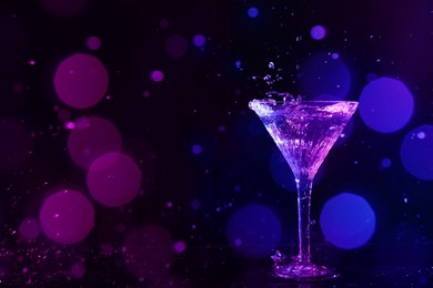 Photo of Glass of martini and splashes in neon lights on dark background, bokeh effect with space for text
