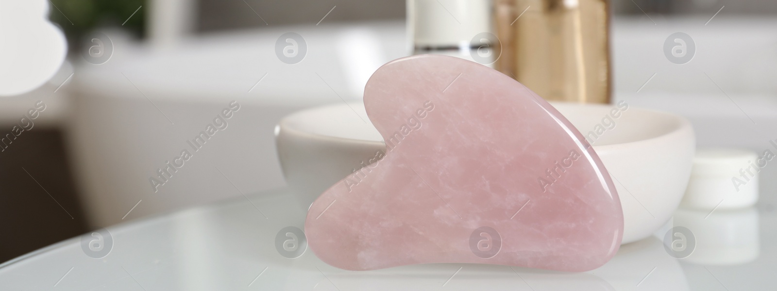 Image of Rose quartz gua sha tool and cosmetic products on white table in bathroom, closeup. Banner design