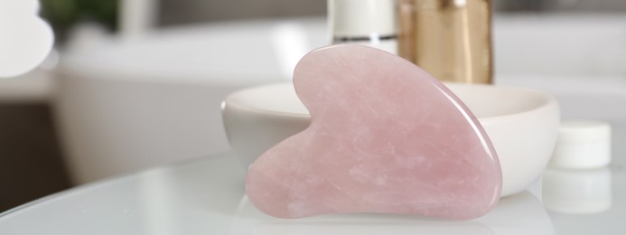 Rose quartz gua sha tool and cosmetic products on white table in bathroom, closeup. Banner design