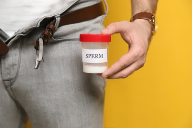 Donor with unzipped pants holding container of sperm on yellow background, closeup