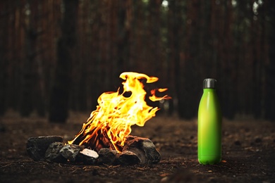 Green thermos bottle near bonfire with burning firewood in forest