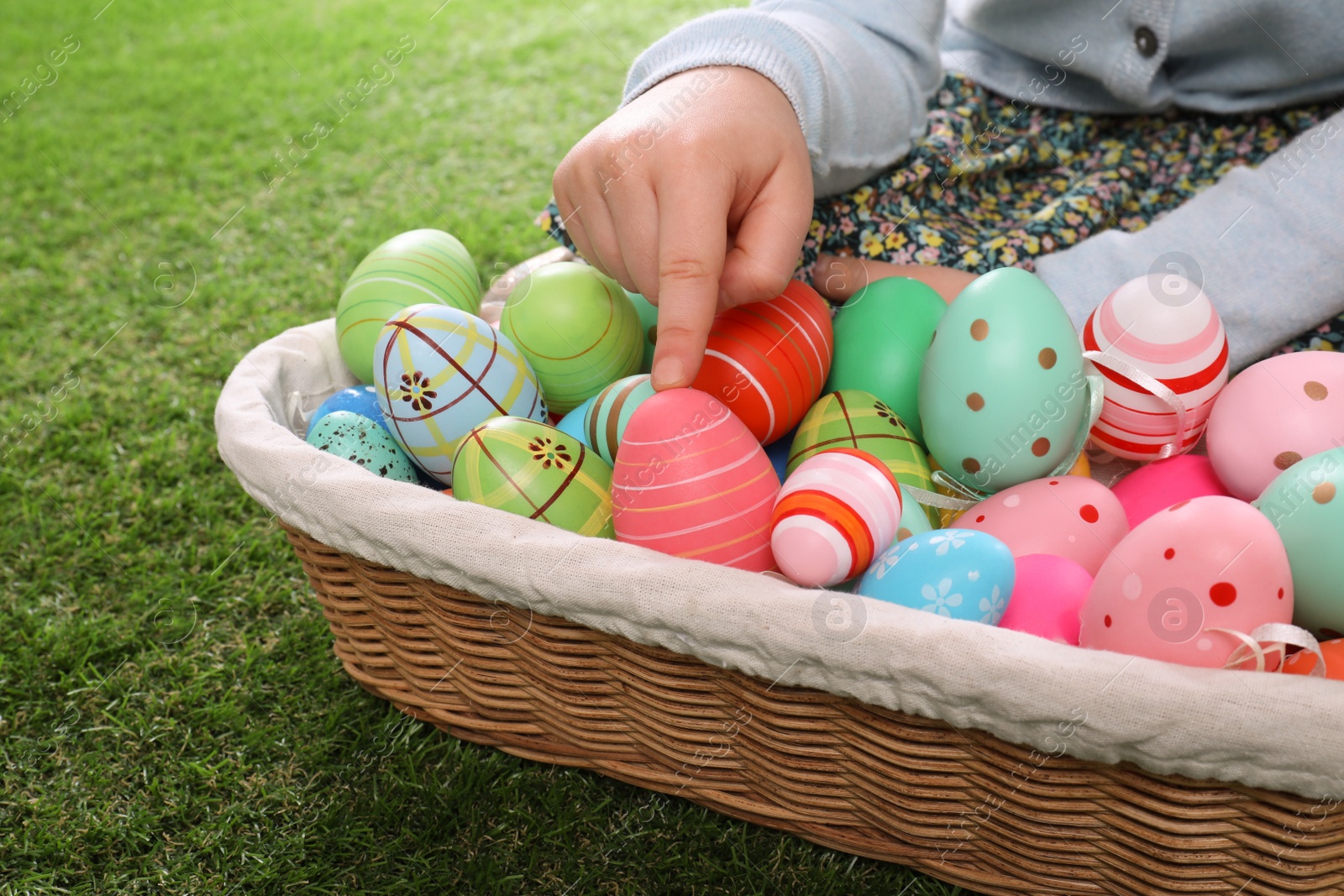 Photo of Child with wicker basket full of Easter eggs on green grass, closeup